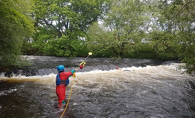 A paddler in a red drysuit, blue bouyancy aid and blue helmet throws a paddle with a rope attached to it, to another person who is stuck in a weir.