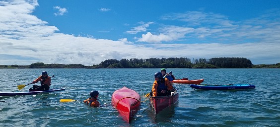 Mae canŵ a phadlwr yn cael eu hachub gan ddau badlwr canŵ arall ar Asesiad Arweinydd Teithio Paddlesport tra bod caiaciwr a phadlfwrdd yn gwylio.